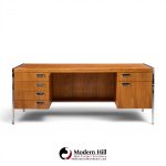 Wooden mid-century desk with metal legs, drawer stacks on both sides, and an open center, set against a white backdrop with branding below.